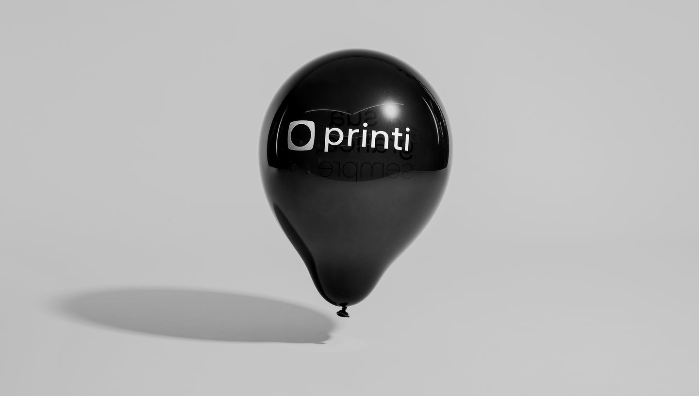 Balão personalizado preto inflado com a marca Printi em destaque, posicionado contra fundo cinza neutro para realce visual.