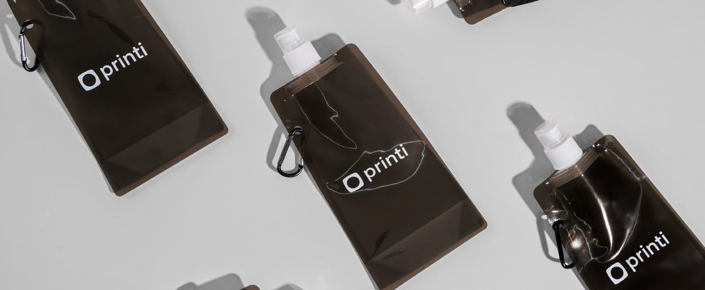 Garrafas flexíveis personalizadas com logo Printi, feitas em material plástico dobrável com tampa tipo bico e mosquetão para prender em mochila, ideais para atividades ao ar livre, eventos esportivos, brindes promocionais e campanhas de marketing sustentá