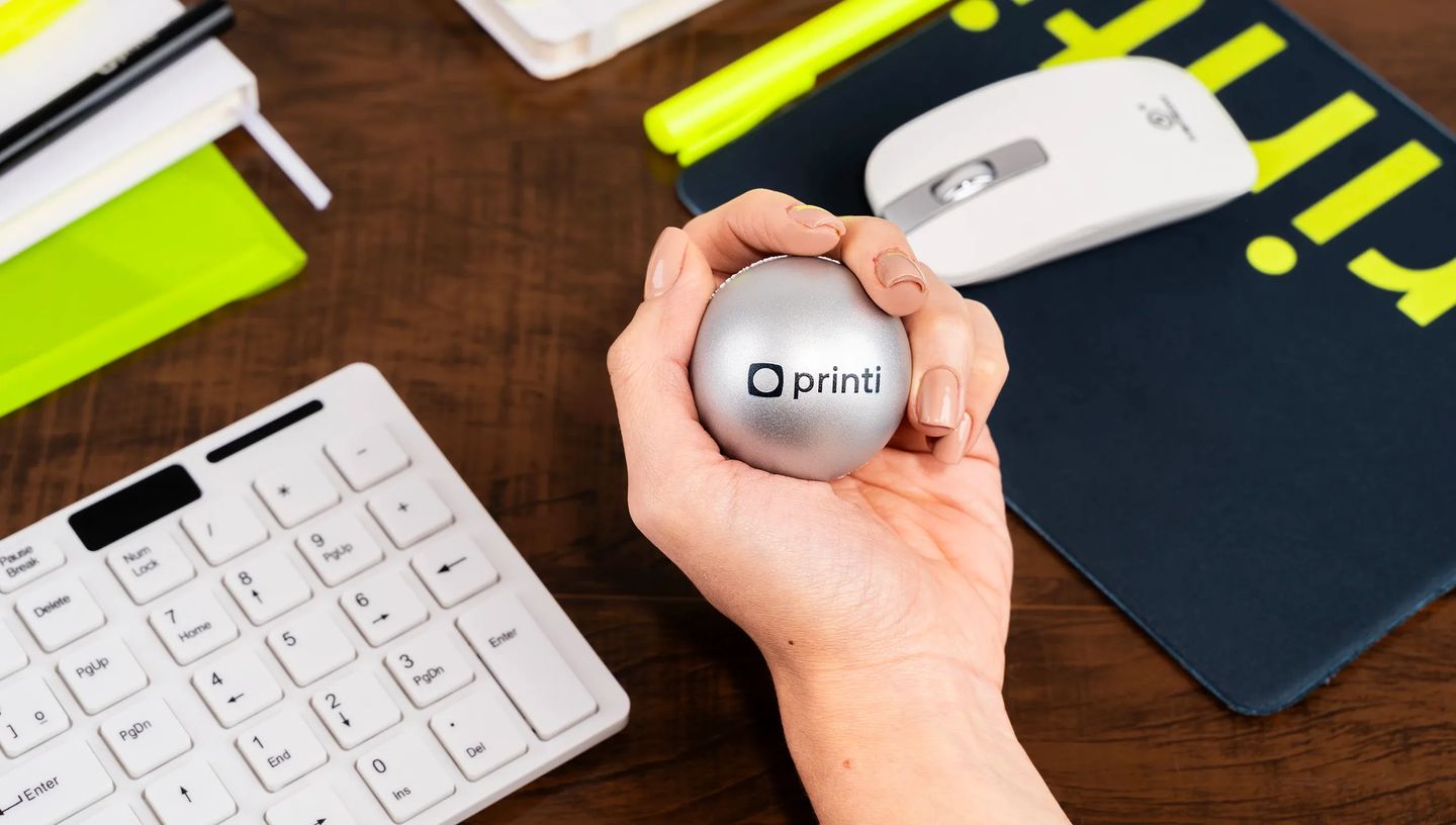 Mão apertando bolinha antiestress prata personalizada com logotipo Printi em ambiente de escritório.