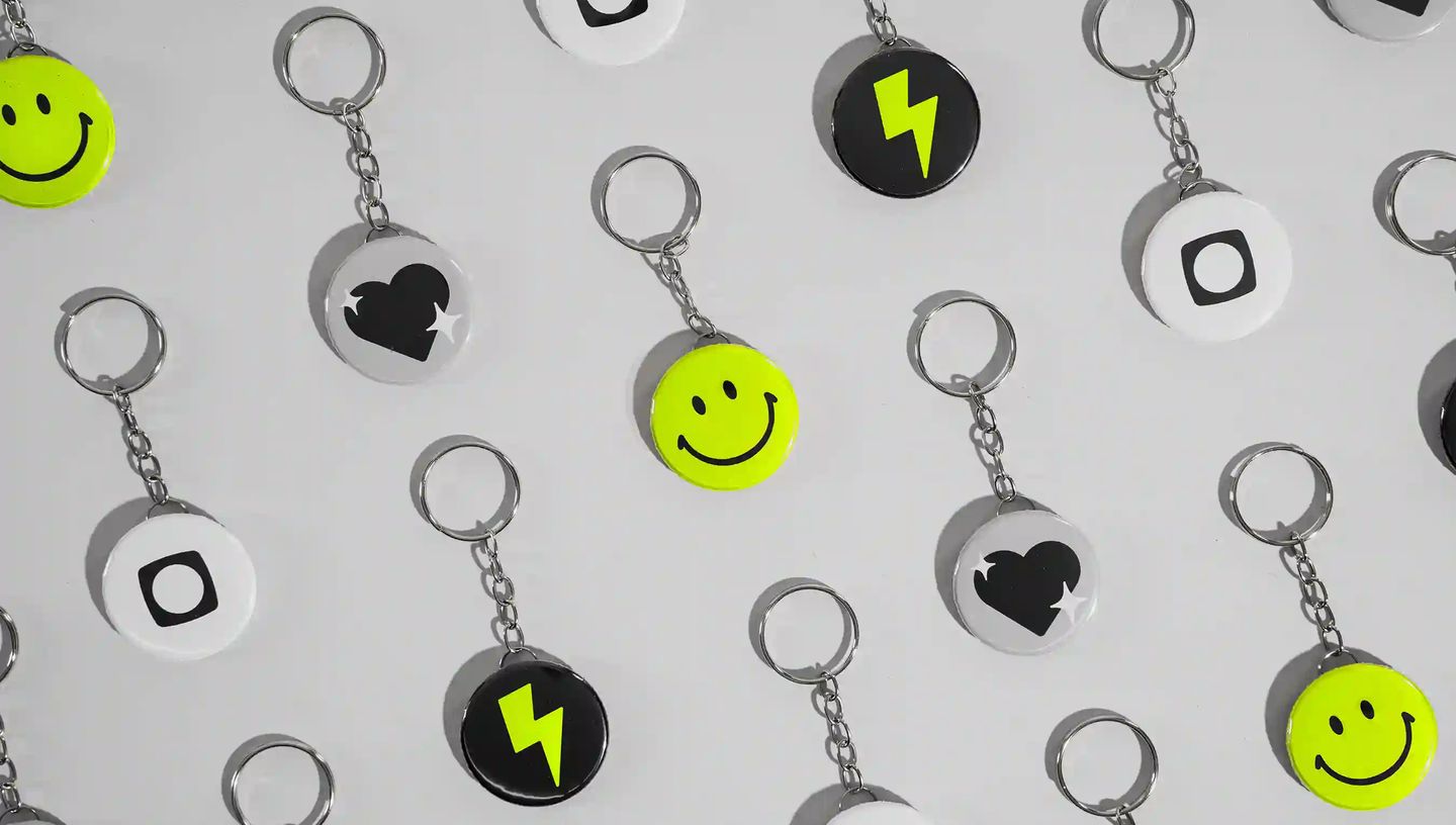 Chaveiros personalizados em formato de botton distribuídos sobre fundo neutro, com designs variados em preto, branco e verde neon, incluindo ícones de sorriso, raio e coração
