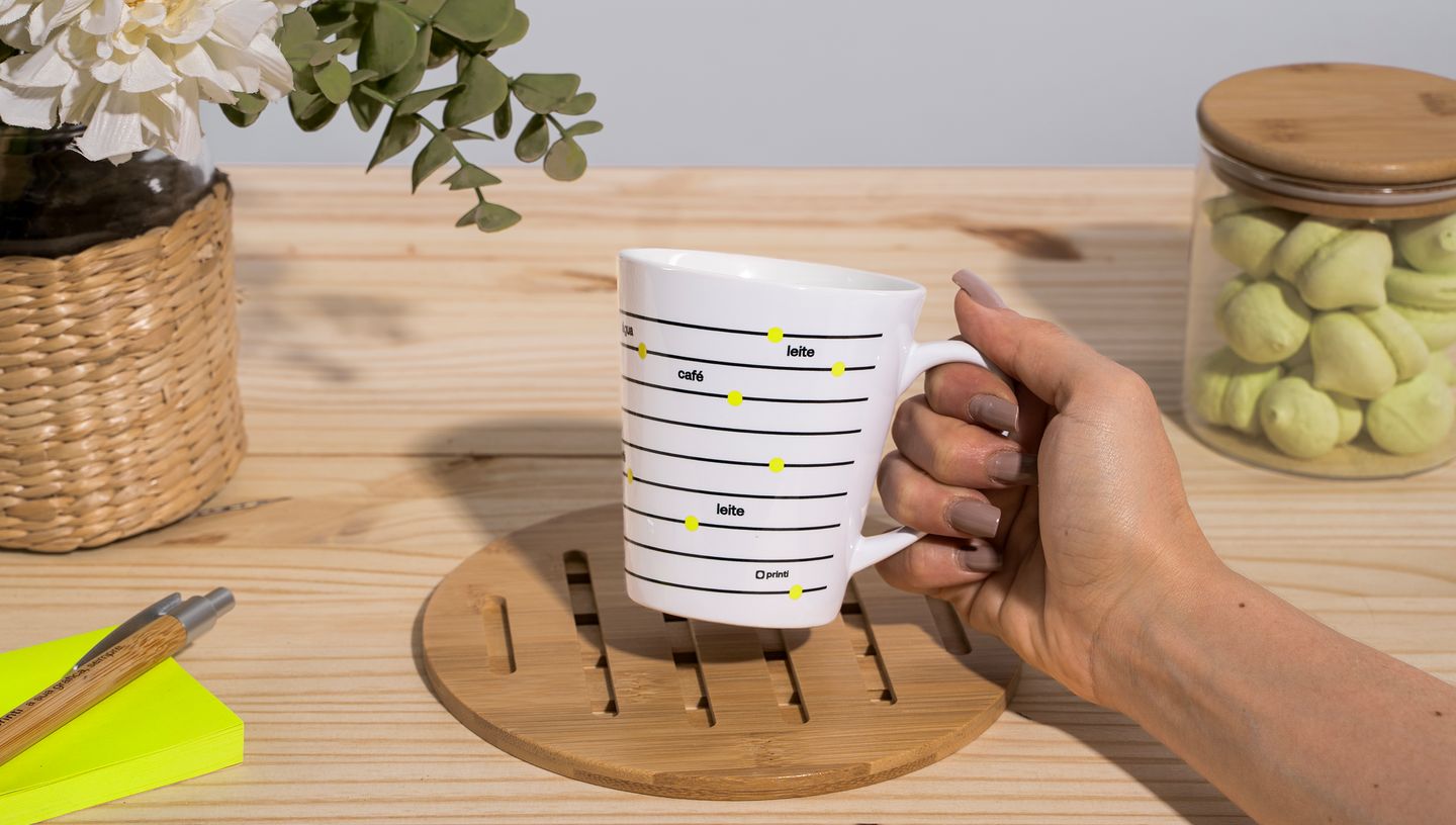 Caneca cônica personalizada sendo segurada sobre uma mesa de madeira, com palavras como café, chá e leite marcadas em linhas horizontais.