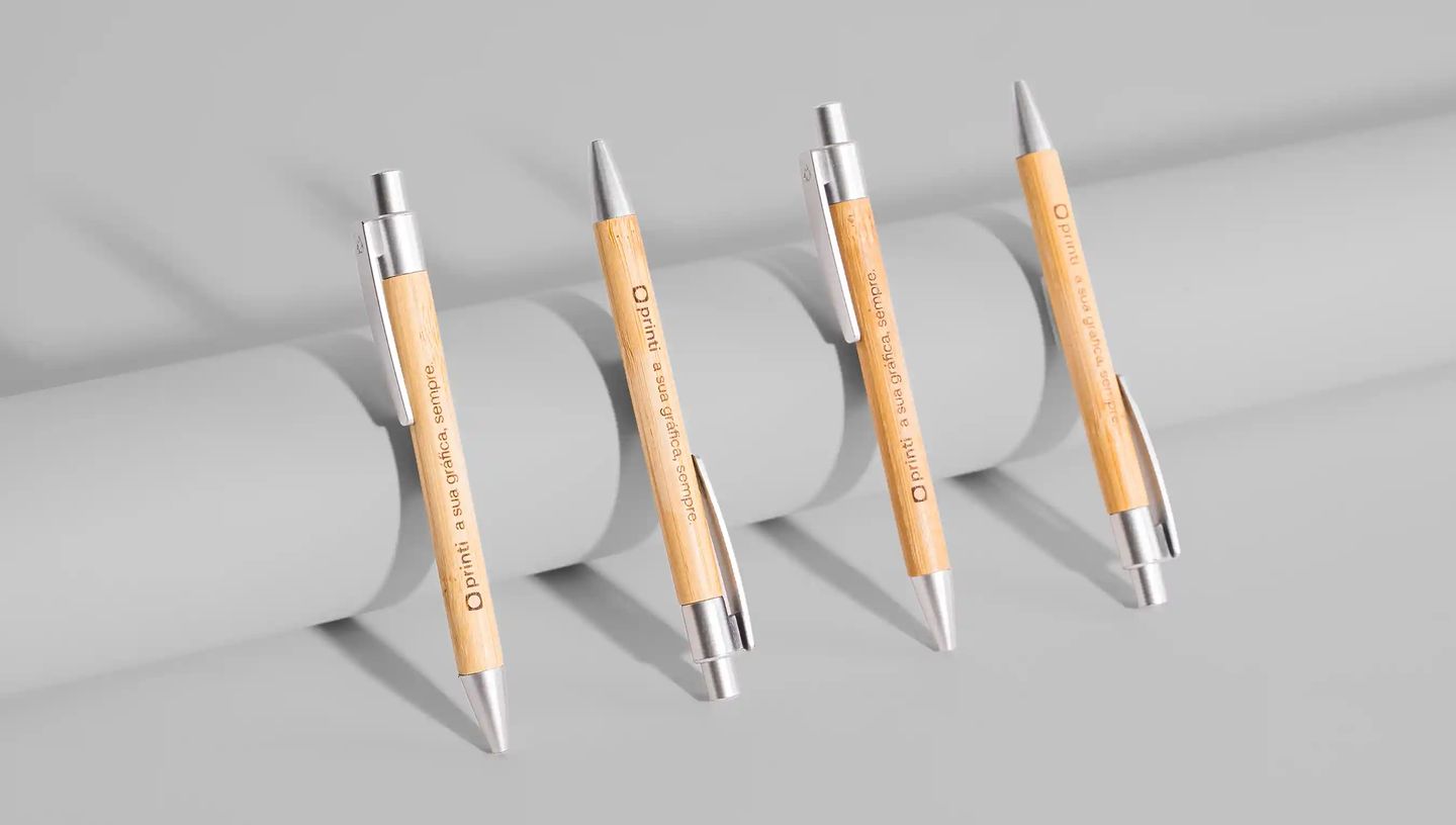 Conjunto de canetas ecológicas de bambu personalizadas com detalhes metálicos prateados e gravação da marca Printi e da frase “A sua gráfica, sempre.” no corpo, dispostas alinhadas sobre fundo neutro, destacando o design sustentável e o uso como brinde co