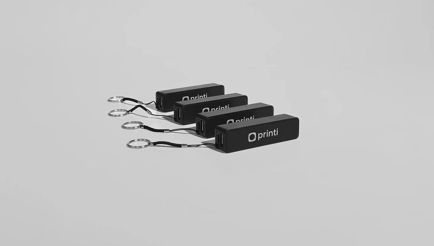 Quatro carregadores portáteis (power banks) personalizados na cor preta com o logo da Printi, alinhados lado a lado sobre fundo neutro e acompanhados de cordões com argolas metálicas para chaveiro, ilustrando um brinde corporativo tecnológico utilizado pa