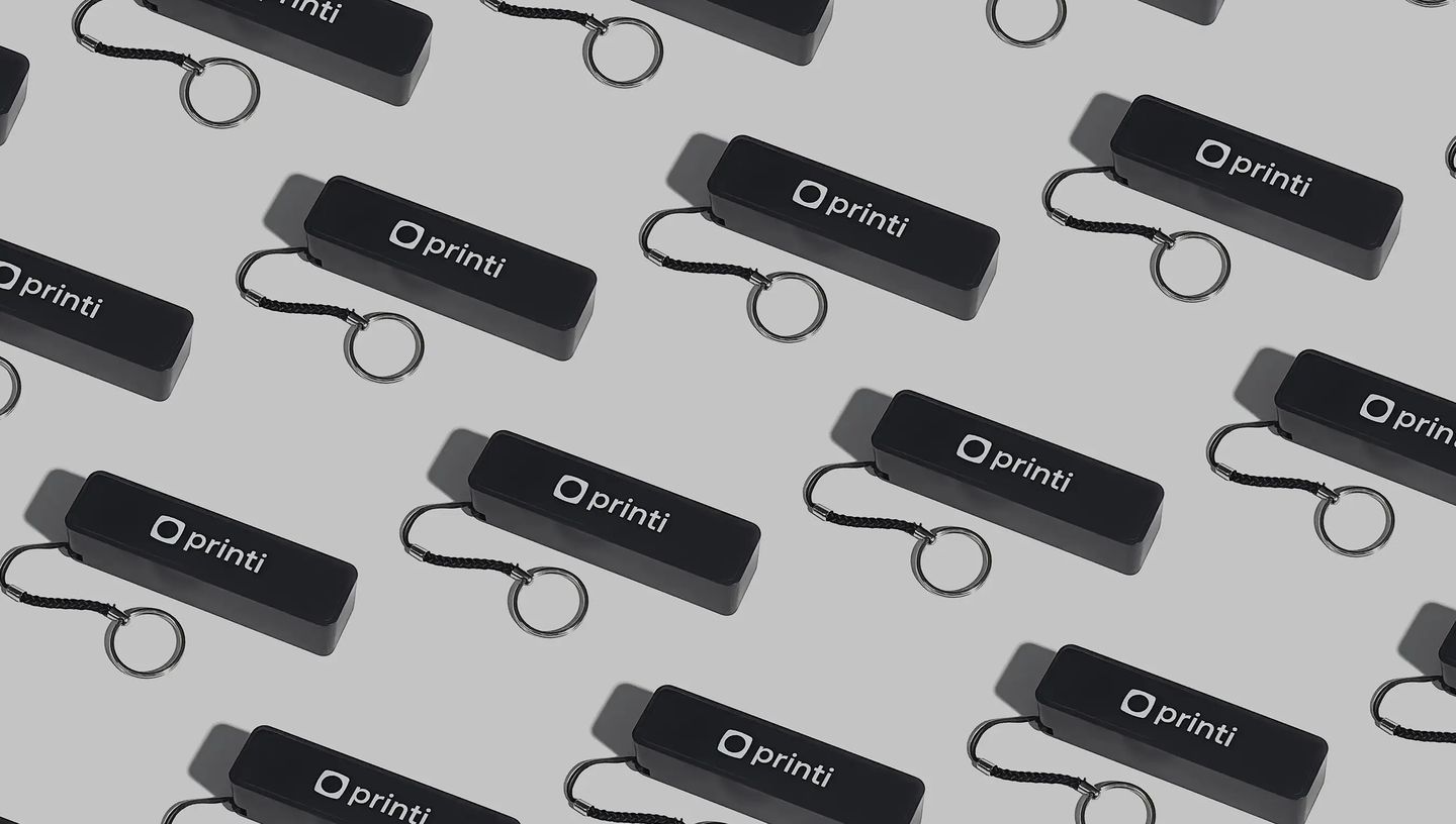 Carregadores portáteis personalizados da Printi na cor preta, com logo em branco e cordão com argola metálica para chaveiro, repetidos em padrão sobre fundo neutro, representando power banks promocionais usados como brindes corporativos para recarregar sm