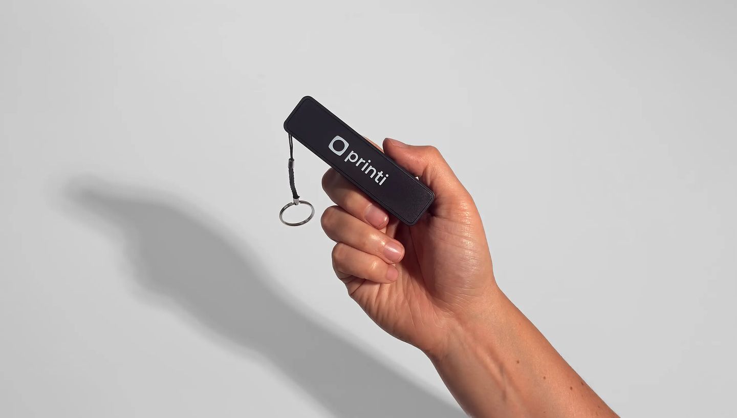 Carregador portátil personalizado da Printi com logotipo branco em corpo preto, segurado por uma mão e com chaveiro metálico, representando brinde corporativo tecnológico ideal para ações promocionais, eventos, kits de onboarding e estratégias de marketin