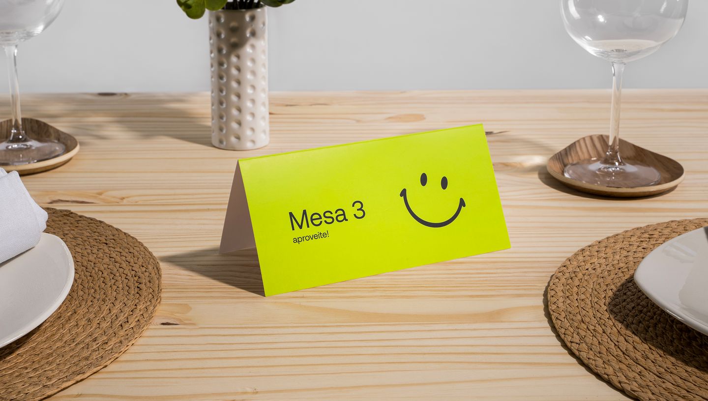 Cartão de mesa posicionado sobre uma mesa posta com jogo americano de palha, pratos e taças, destacando-se pelo design moderno e alegre na cor amarela neon, com o texto “Mesa 3, aproveite!”