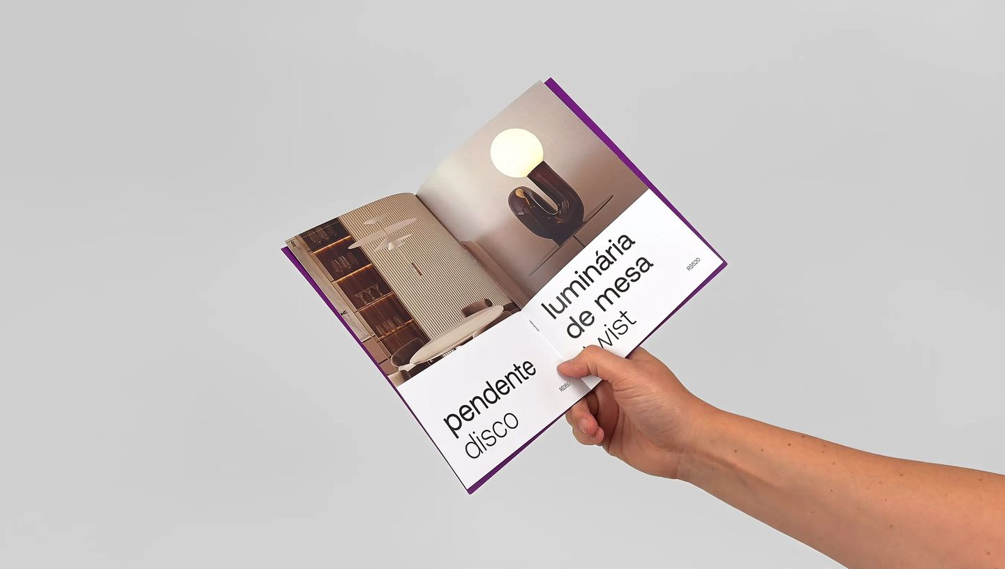 Catálogo personalizado impresso sendo segurado por uma mão, aberto em página com fotos de luminária e design editorial moderno.