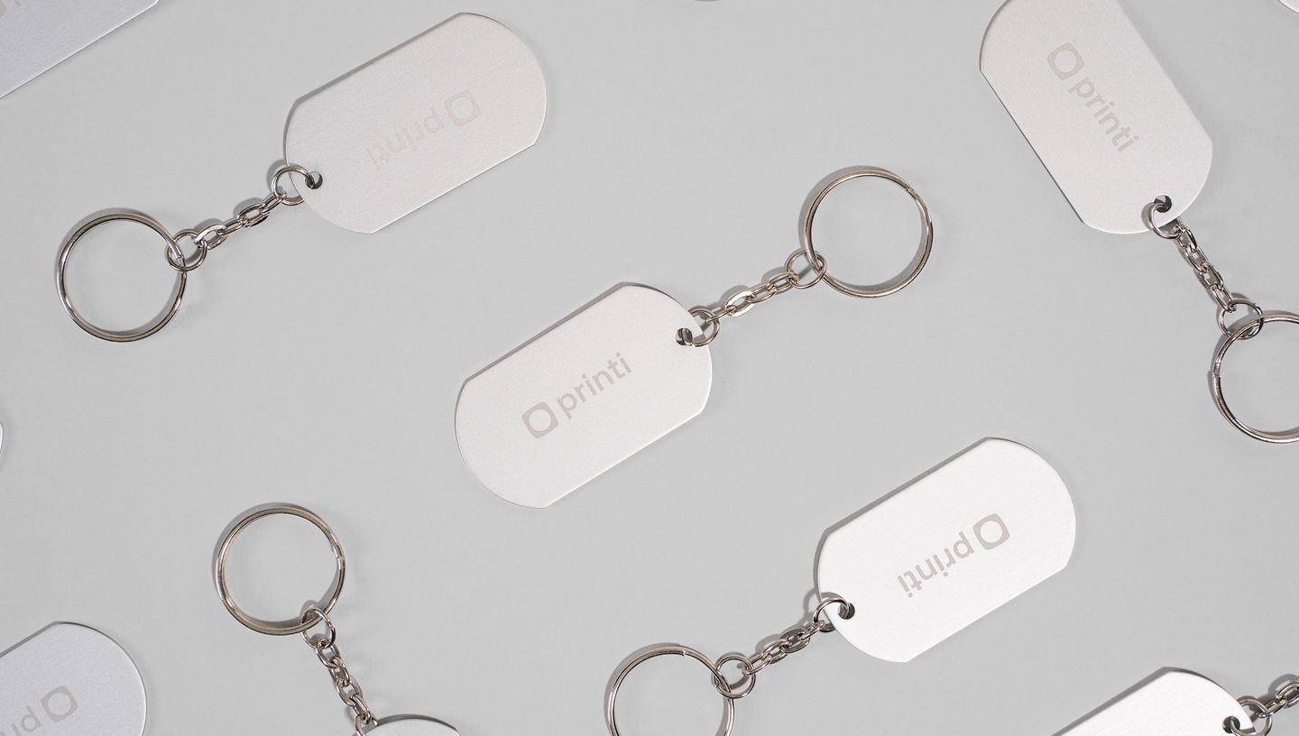 Diversos Chaveiros de Metal Básico com plaquinha retangular prateada personalizada com logo da Printi, exibido em padrão repetido sobre fundo cinza claro