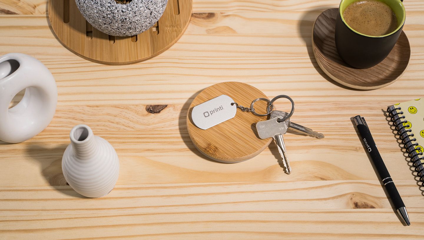 Chaveiro Metal Básico com plaquinha de alumínio personalizada com logo da Printi, apoiado sobre porta-chave de bambu em mesa de madeira clara com objetos decorativos ao redor