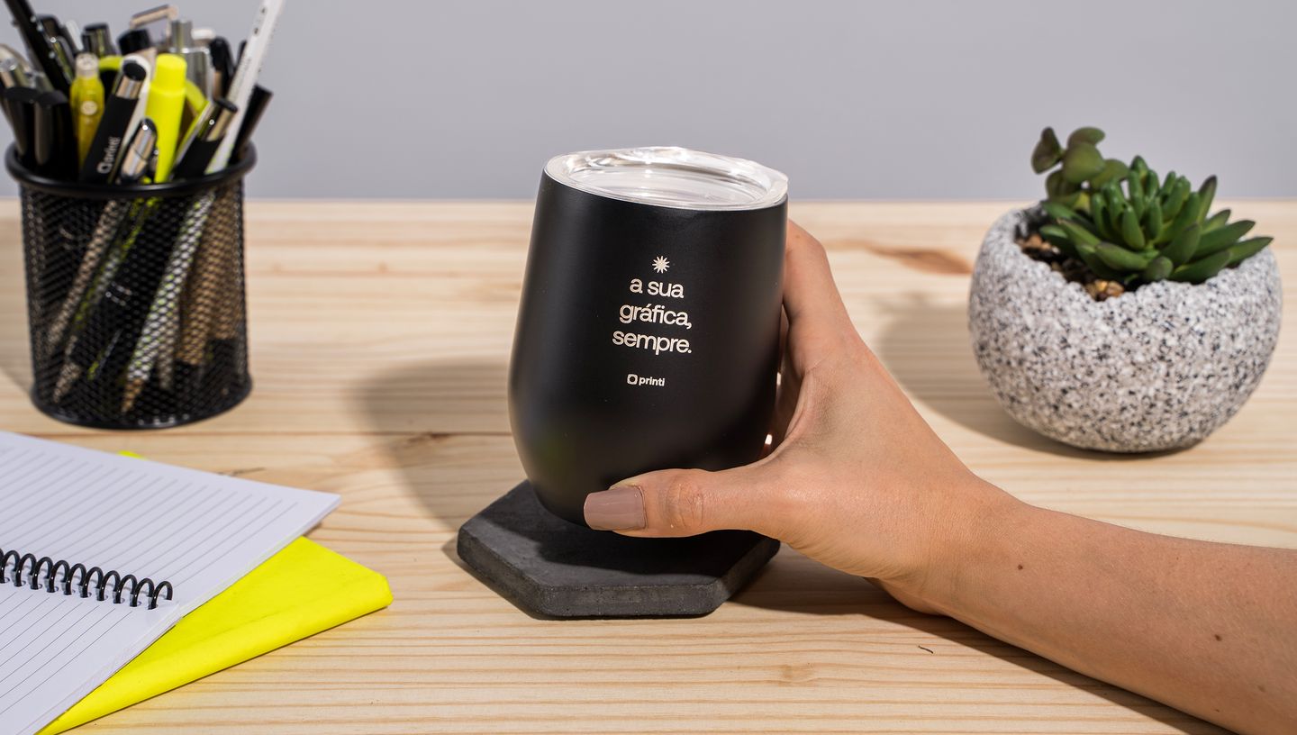 Mão de uma pessoa branca apoiada em uma superfície de madeira segurando um Copo Térmico de 400 ml preto com tampa, personalizado com a frase "a sua gráfica, sempre", com um papel branco e amarelo ao lado, um porta lápis com diversas canetas e lápis dentro