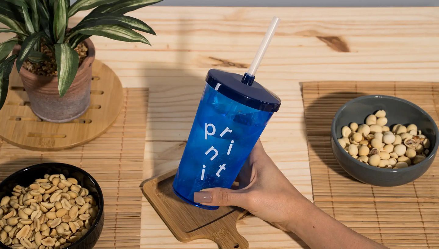 Copo twister personalizado azul translúcido com tampa e canudo, exibindo a marca Printi, sendo segurado por uma pessoa sobre uma mesa de madeira clara. Ao redor do copo aparecem tigelas com amendoins e outros petiscos, além de uma planta decorativa em vas
