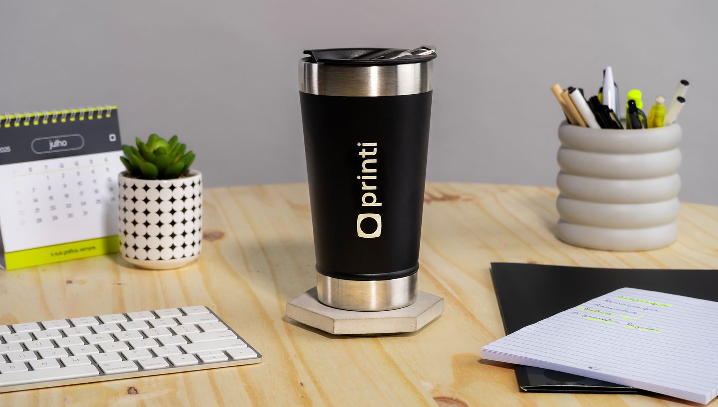 Copo térmico com tampa sobre uma mesa de madeira de escritório, ao lado de um teclado, bloco de notas, calendário, porta lápis e um vasinho branco com planta