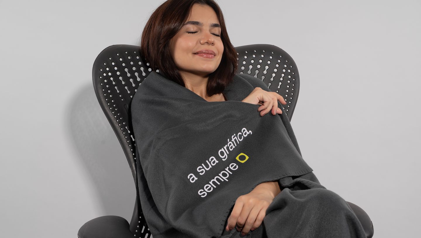 Mulher sentada em cadeira utilizando a manta polar preta com slogan da Printi