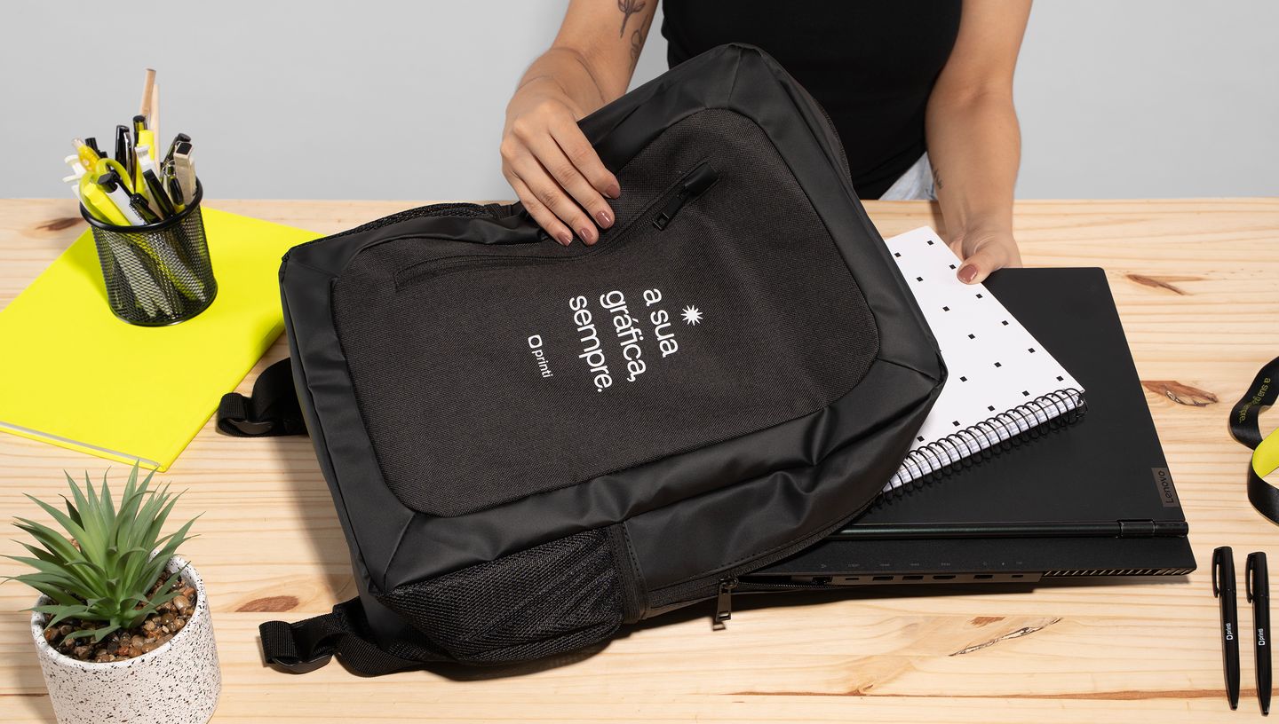 Pessoa colocando um caderno dentro de uma mochila preta sobre uma mesa com itens de papelaria e uma suculenta.