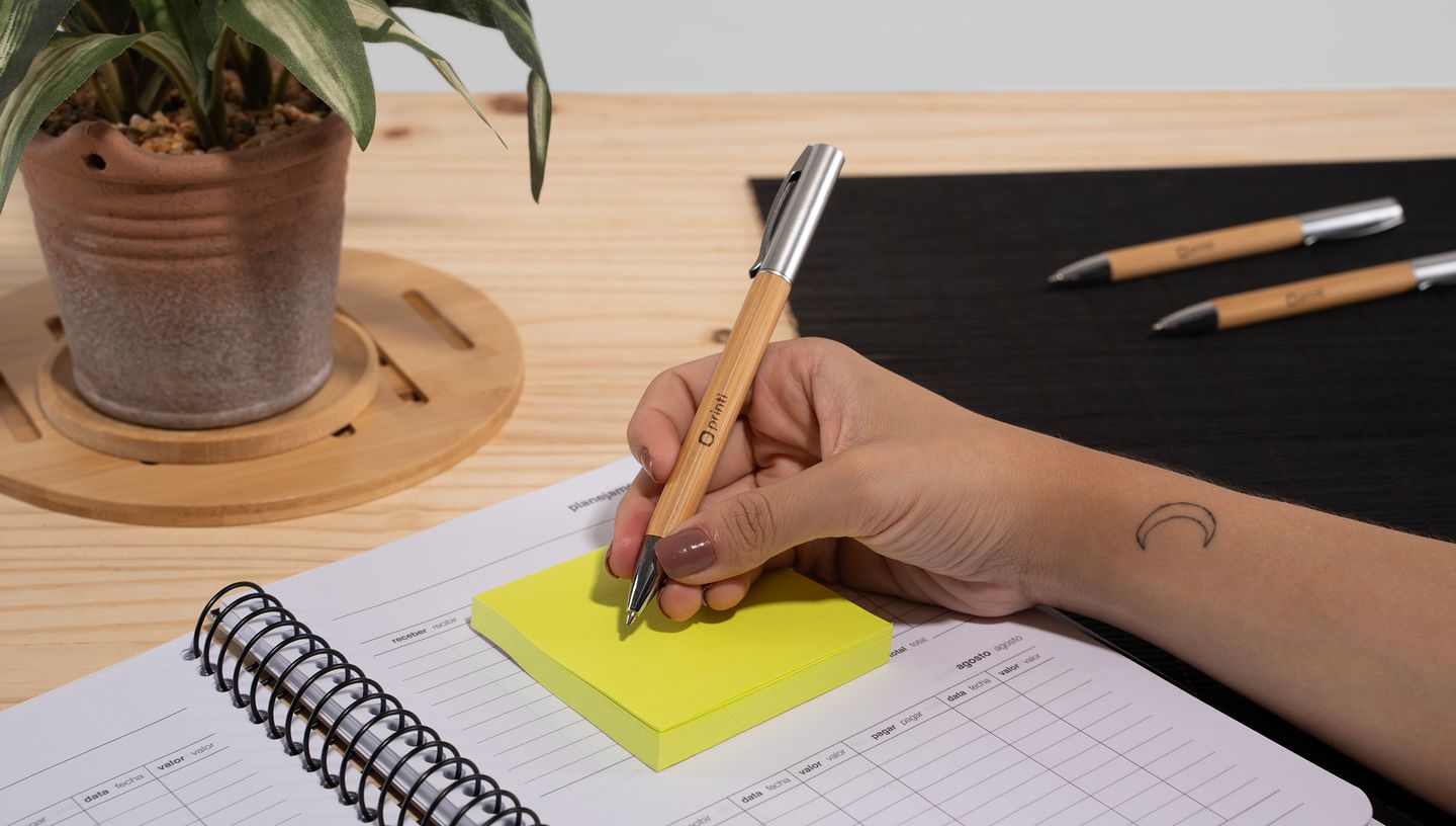 Pessoa com caneta em bambu beal na mão escrevendo em bloco de notas, em cima de caderno sobre mesa de madeira com vaso de planta e duas canetas perto..