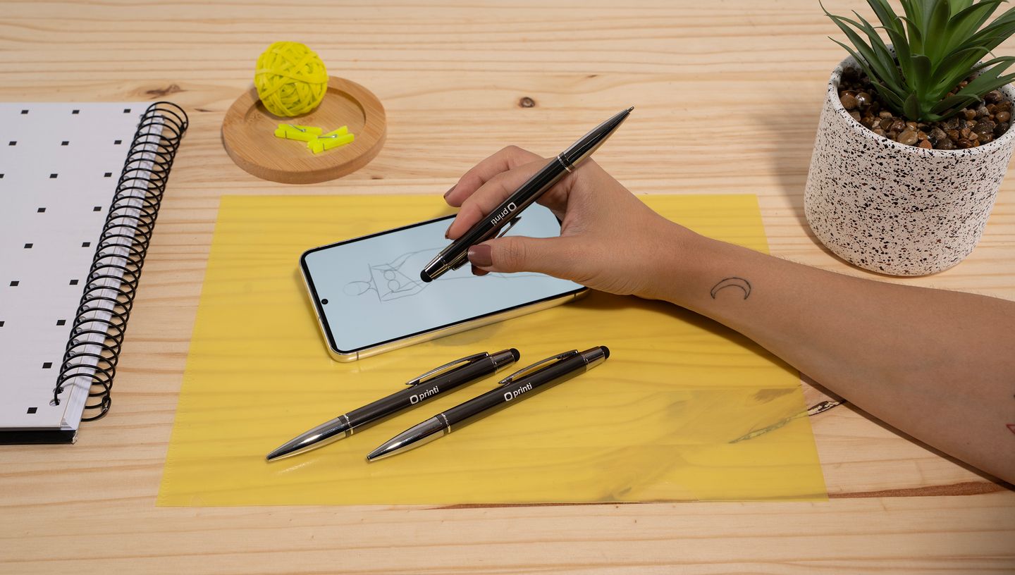 Pessoa utilizando uma caneta touch em um smartphone sobre uma mesa de madeira com bloco de notas, outras canetas, planta decorativa e itens de papelaria.