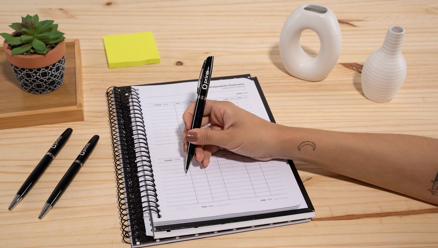 Pessoa escrevendo em caderno com caneta de metal lando personalizada na cor preta sobre mesa de madeira.