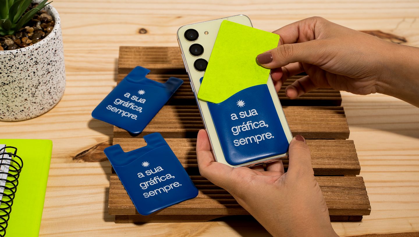 Pessoa segurando celular com porta cartão na cor azul e cartão na cor verde, em mesa de madeira com outros porta cartões.