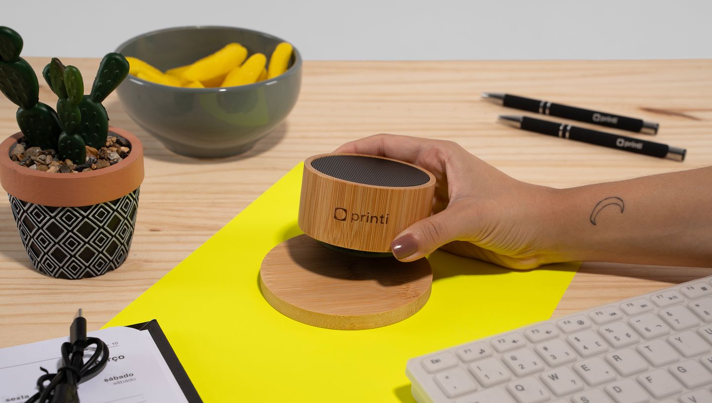 Pessoa segurando caixa de som em bambu personalizada em mesa com objetos e planta ao redor.