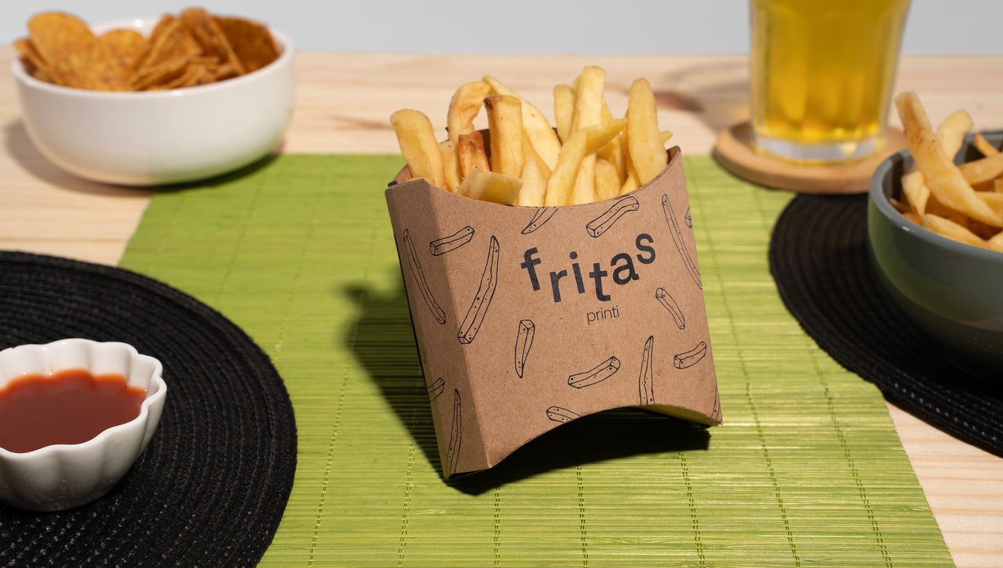 Batata frita em embalagem de papel sobre jogo americano verde; ao redor, há potinho com molho, tigela com chips, copo de bebida e outra porção de fritas.