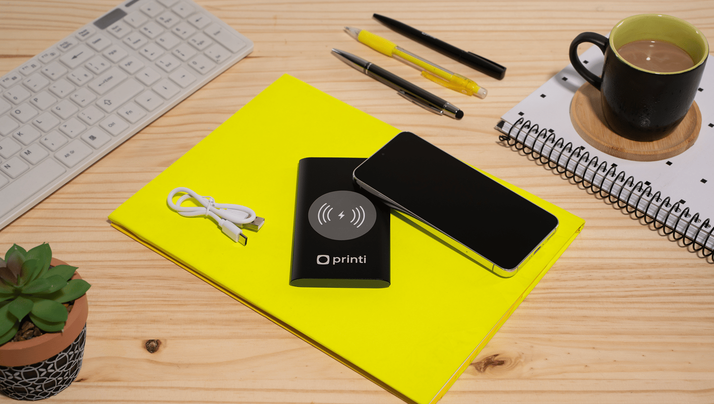 Carregador portátil por indução com celular em cima de objeto amarelo em mesa de madeira com suculenta, canetas e xicará com café e teclado ao redor