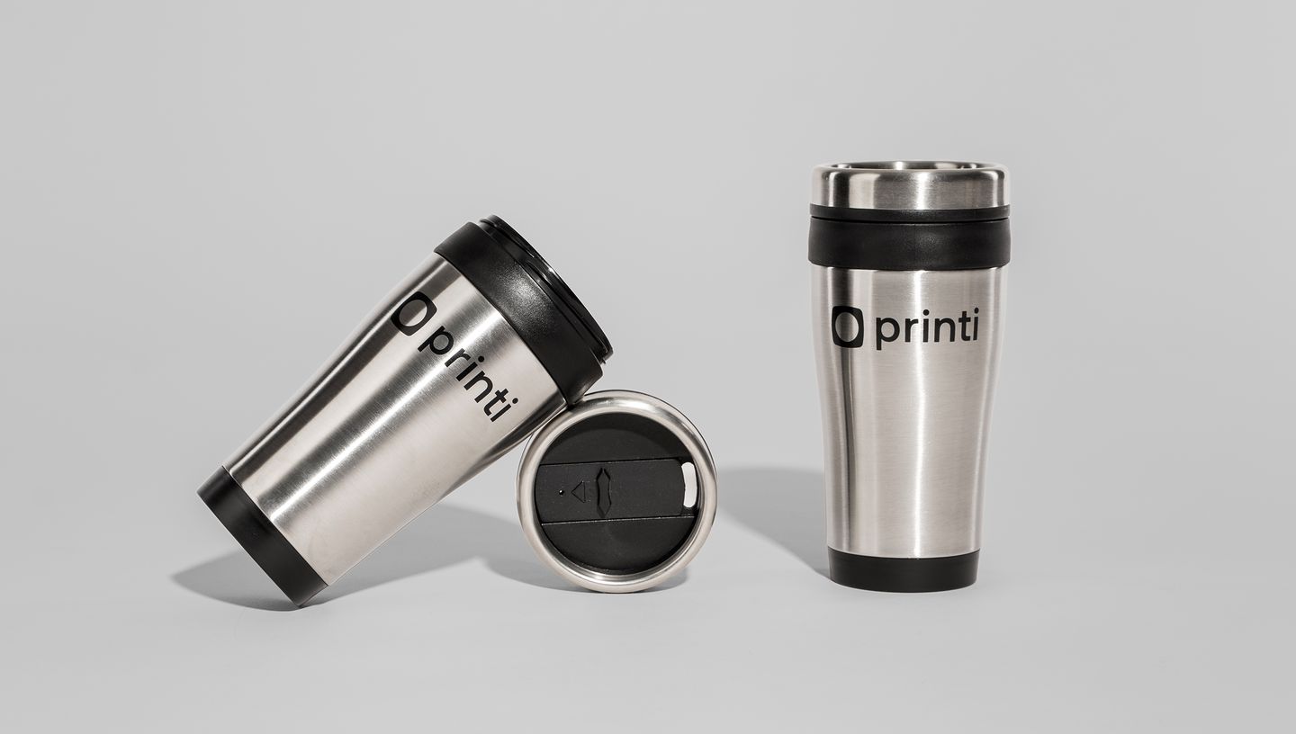 Três copos térmicos de inox com tampa preta em exibição sobre fundo cinza claro — um copo térmico deitado com o logotipo Printi visível, outro exibindo a tampa com abertura deslizante, e o terceiro em pé