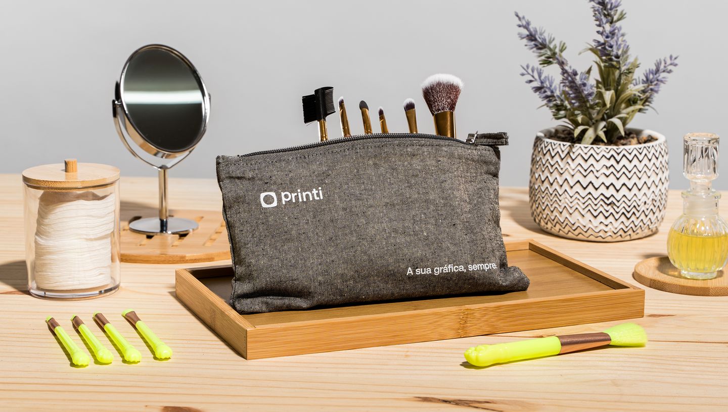 Necessaire personalizada Millie em tecido escuro, com zíper e logo da Printi, exposta com pincéis de maquiagem e acompanhada por espelho, lavanda decorativa, perfume e utensílios de beleza.