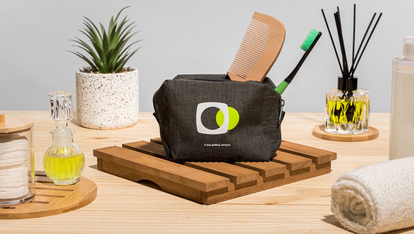 Necessaire preta com estampa personalizada apoiada em base de madeira, cercada por itens de cuidados pessoais como escova, toalha, óleos aromáticos, difusor e planta decorativa.