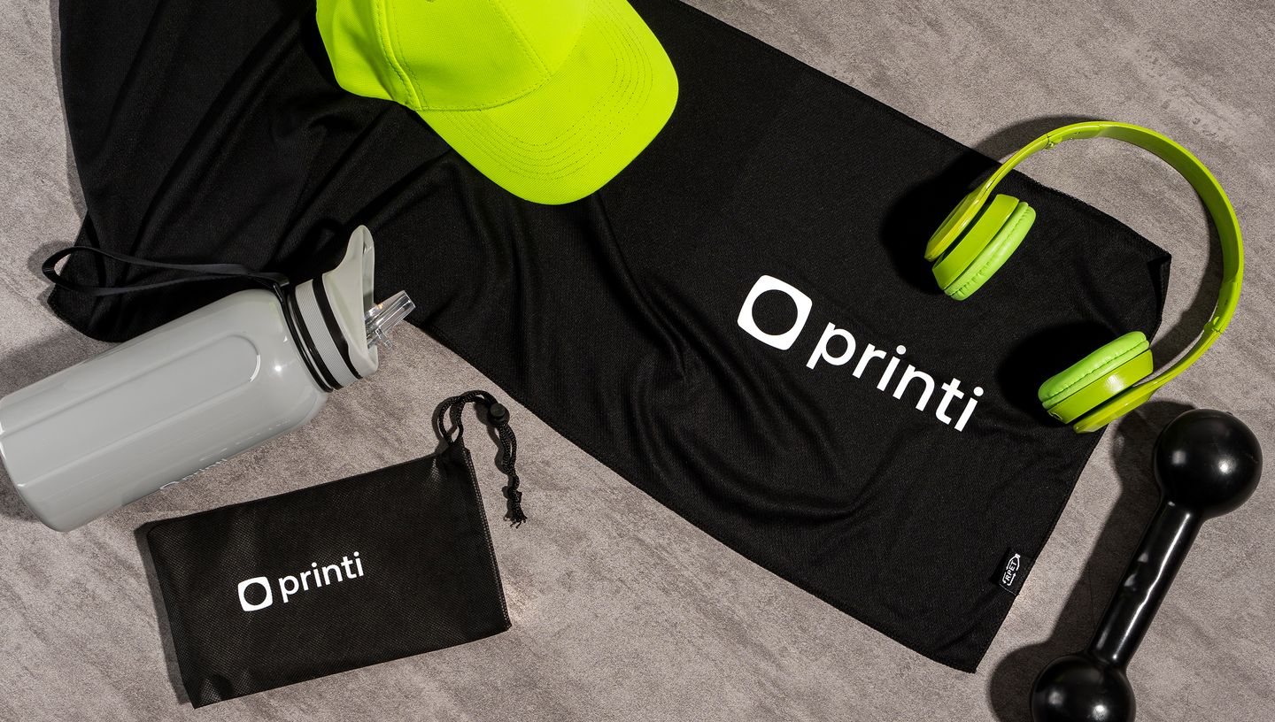 Toalha esportiva em PET com bolsa, ambas na cor preta com logo da Printi ao lado de fone de ouvido, haltere, boné e garrafa de água.