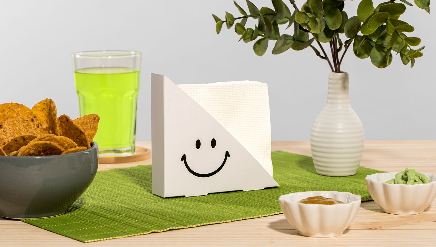 Porta-guardanapo branco com estampa de smiley, apoiado sobre jogo americano verde, ao lado de tigelas com petiscos, bebida verde e vaso decorativo.