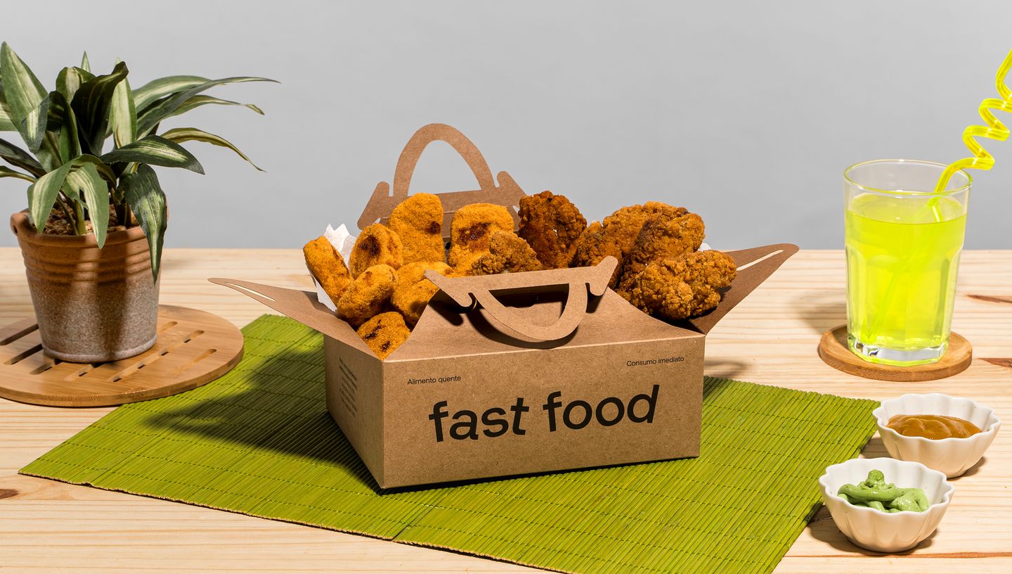 Embalagem para frituras com o nome fast food com frituras dentro, em mesa com vaso de planta e copo com bebida e duas tigelas ao redor. 