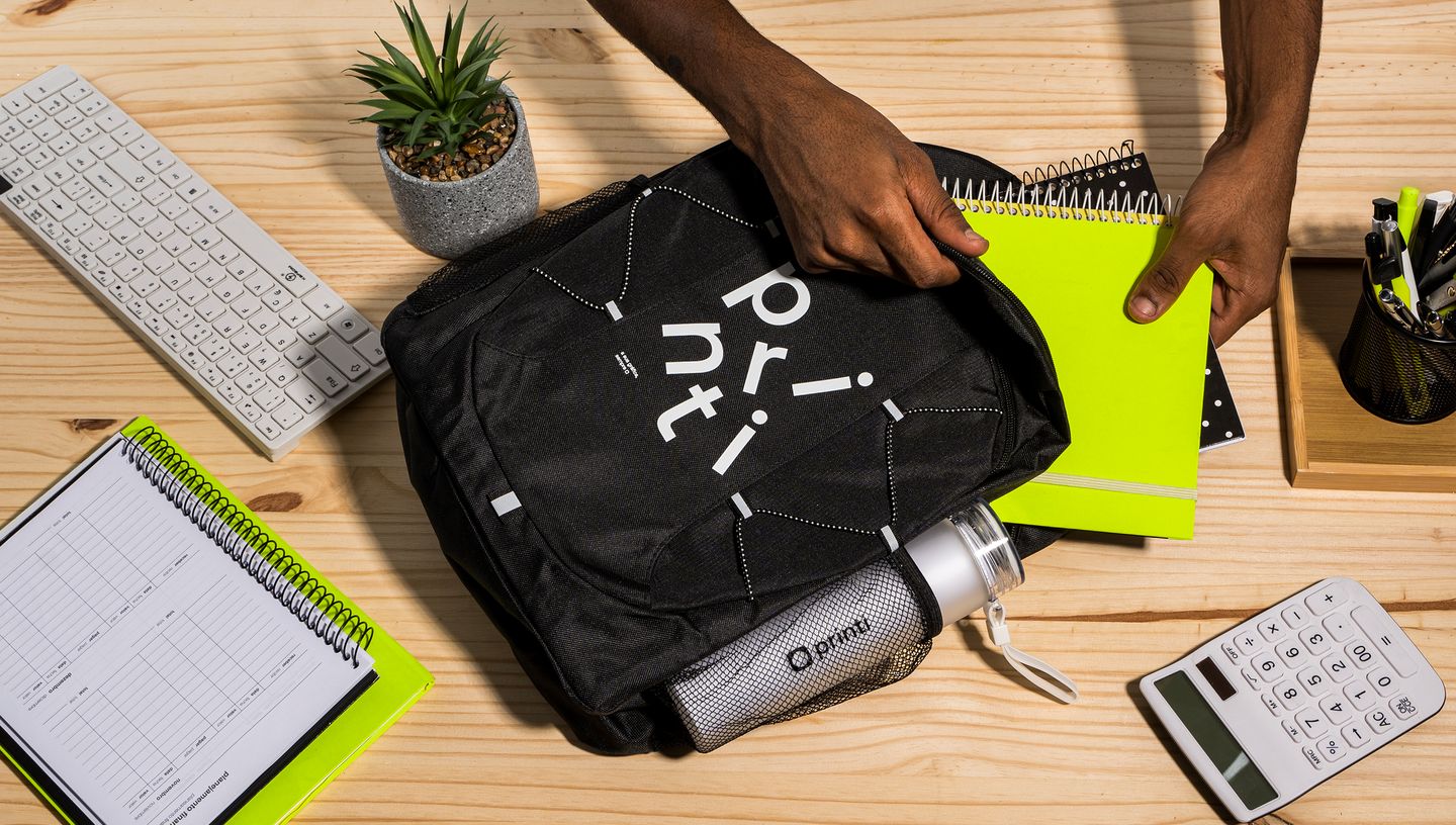 Pessoa colocando caderno dentro de mochila básica com garrafa, com caderno, teclado, suculenta, calculadora e kit de escrita ao redor.