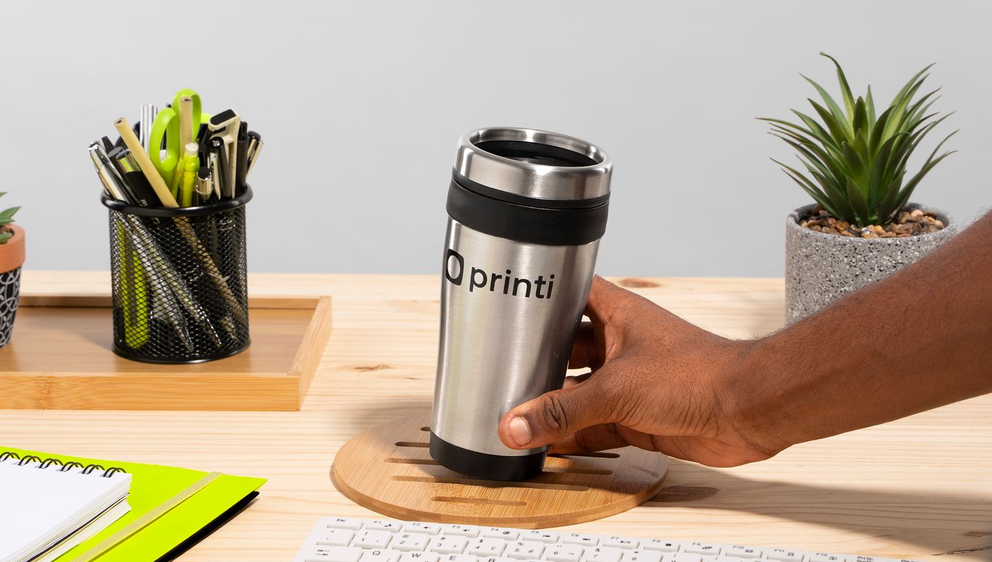 Copo térmico em aço inox com tampa preta sendo segurado por uma mão sobre uma mesa de madeira, rodeado por itens de escritório como caderno, teclado, porta-canetas e vasos decorativos