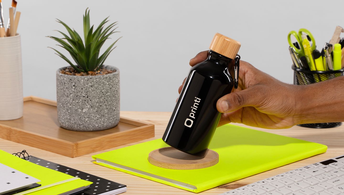Pessoa posicionando um squeeze preto de alumínio reciclado com tampa de madeira e logo da Printi sobre um caderno em uma mesa de escritório.
