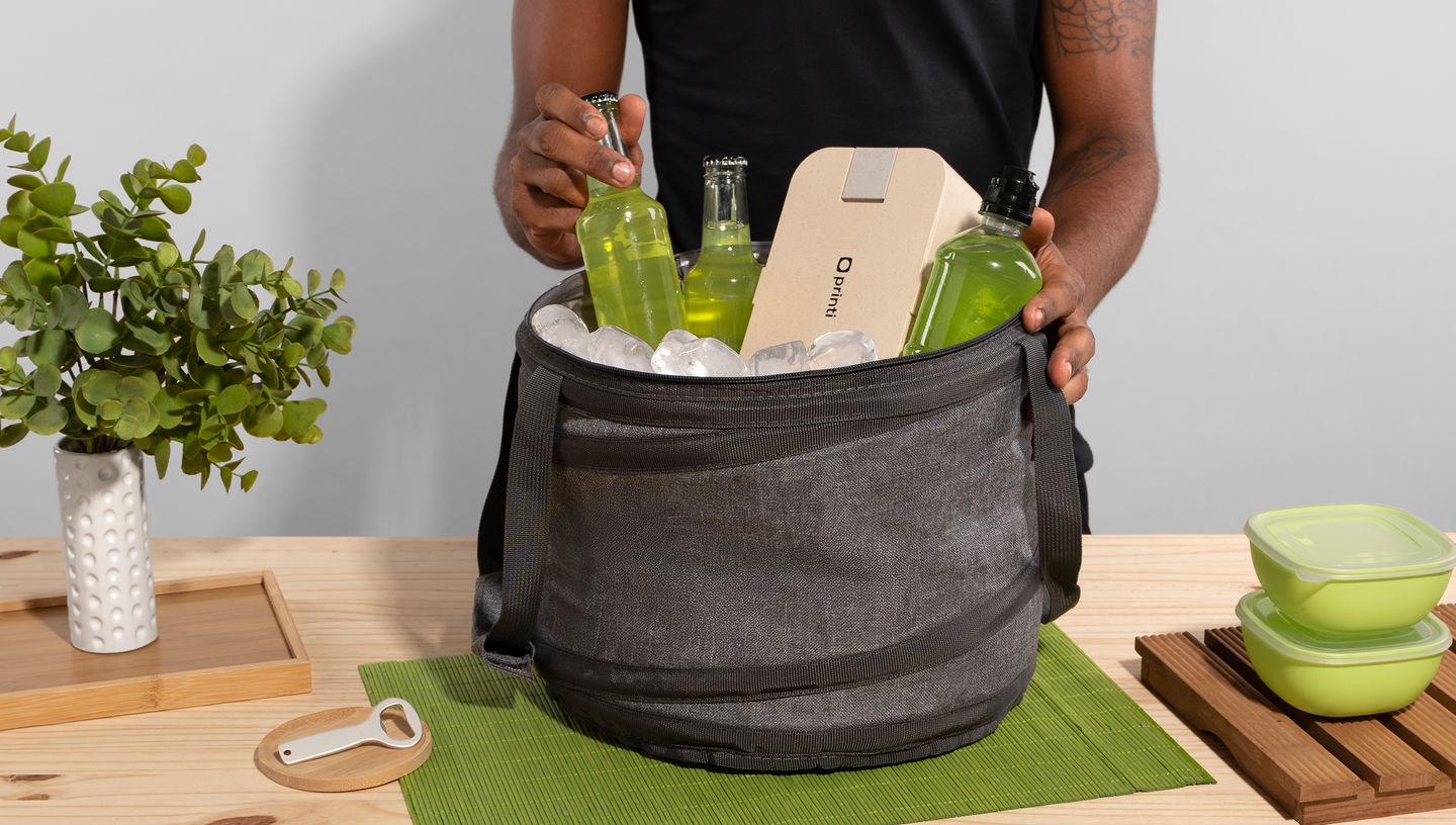 Pessoa colocando uma garrafa de bebida dentro de uma bolsa térmica cinza cheia de gelo em mesa de madeira com planta e coisas de cozinha ao redor.