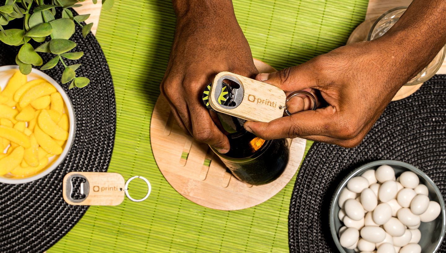 Pessoa usando o chaveiro abridor de bambu personalizado para abrir garrafa de Cerveja com design em madeira, sobre mesa com petiscos e toalha verde.