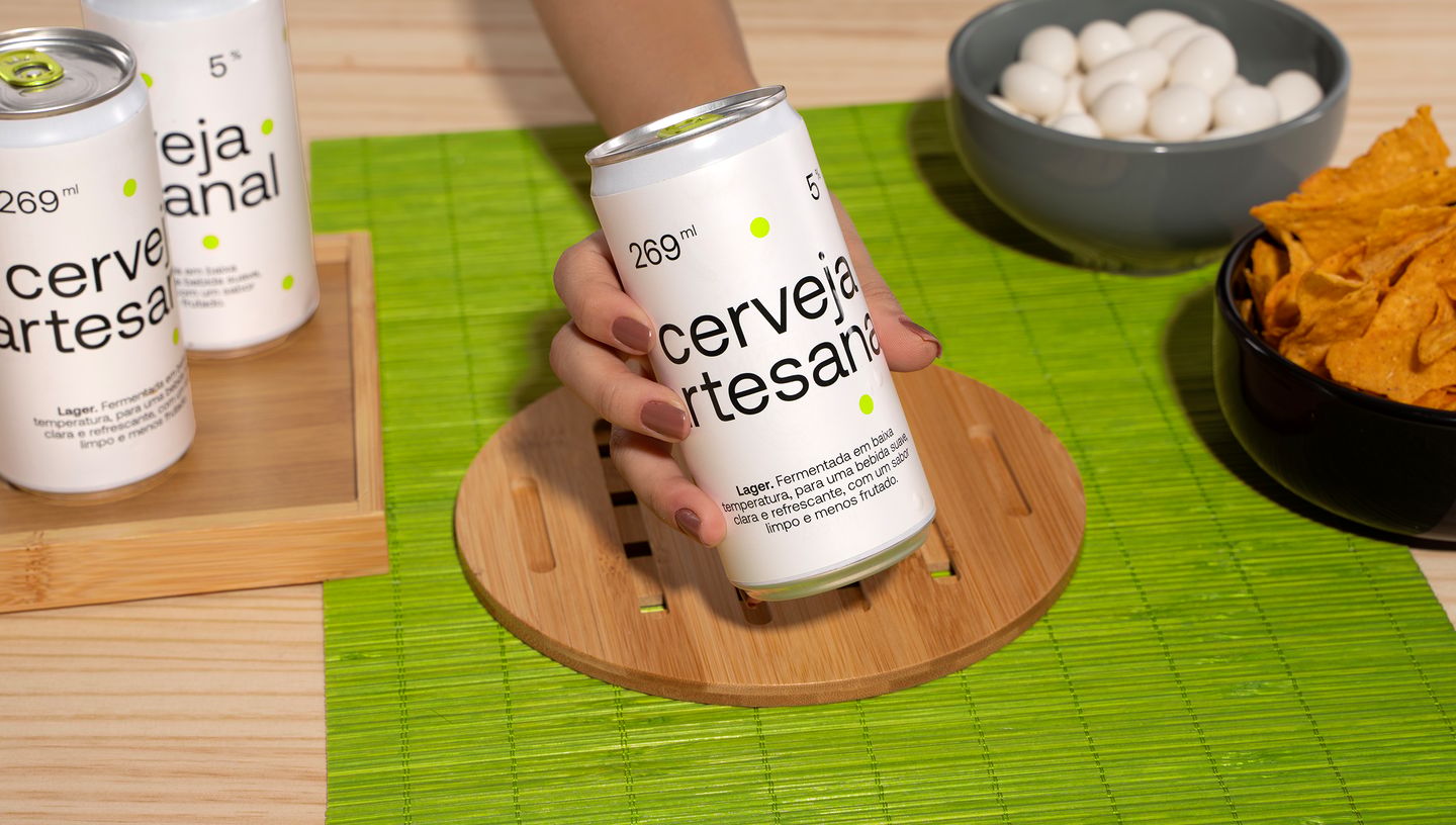 Rótulo para lata personalizado em lata de cerveja com design minimalista em branco, sendo exibido por uma mão sobre suporte de madeira, ao lado de petiscos sobre toalha verde.