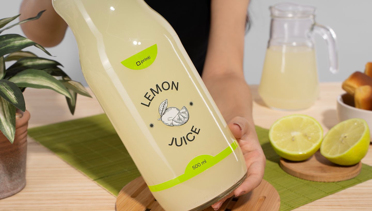 Rótulo para garrafa de suco aplicado em embalagem transparente com o texto “Lemon Juice”, sendo exibido ao lado de limões cortados, jarra e mesa posta.