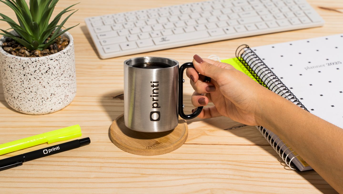 Pessoa segurando caneca para transporte sobre mesa com planta e cadernos ao redor.