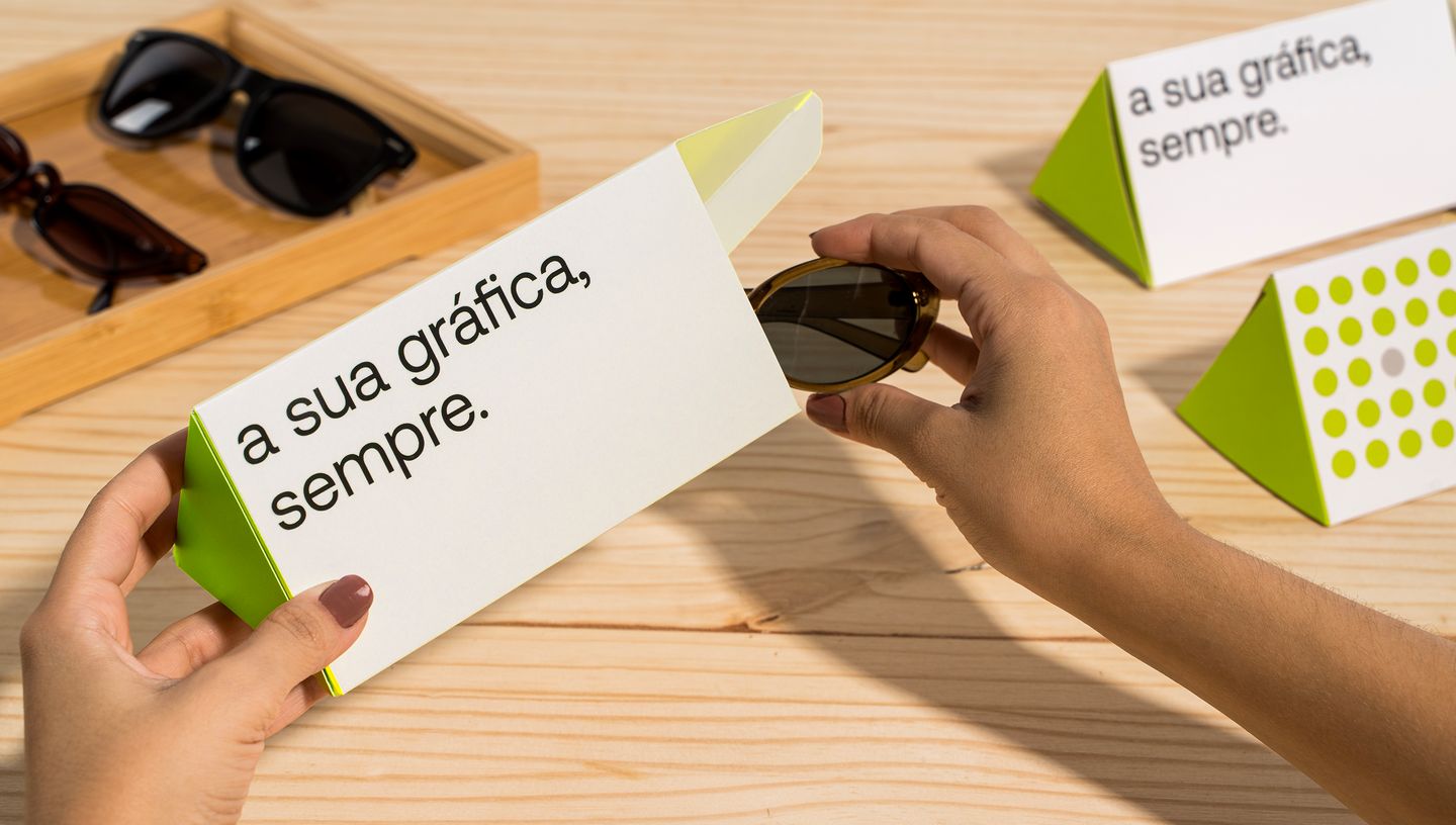 Pessoa colocando óculos dentro de embalagem triangular nas cores branco e verde, com slogam da Printi, além de outras embalagens e óculos no fundo.