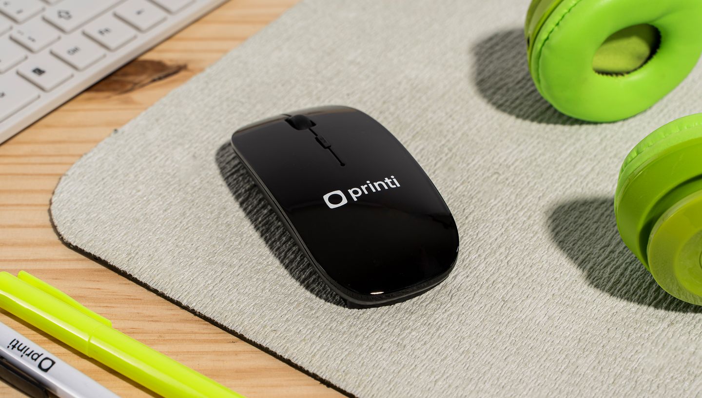 Mouse sem fio personalizado na cor preta com logo da Printi sobre mouse pad, ao lado de fone de ouvido verde.