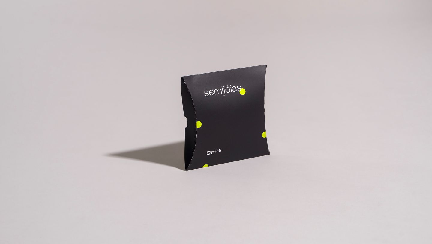 Embalagem almofada bijuteria preta com design moderno e logo Printi, em fundo cinza neutro