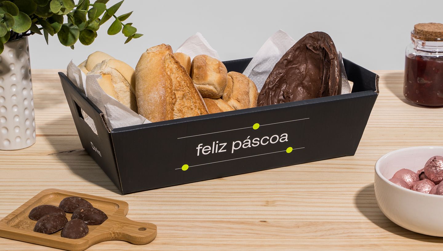 Embalagem cesta preta personalizada com texto escrito feliz pascoa, montada e decorada com pães e chocolates, e apoiada em cima de uma superfície de madeira  e com potes ao redor compondo a decoração