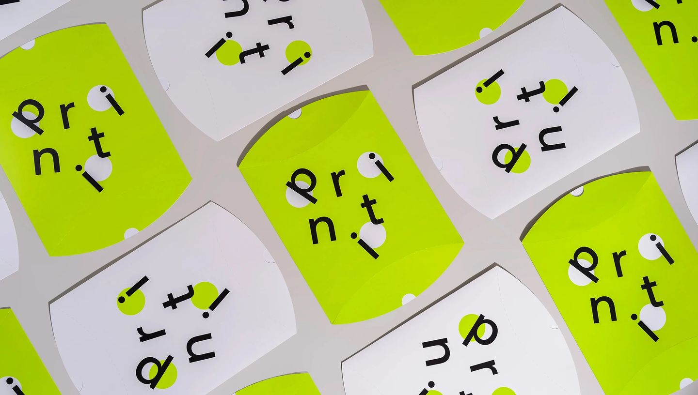 Embalagens tipo almofada personalizadas em papel cartão nas cores branco e verde neon com a marca Printi impressa em preto, dispostas em padrão repetido sobre fundo neutro