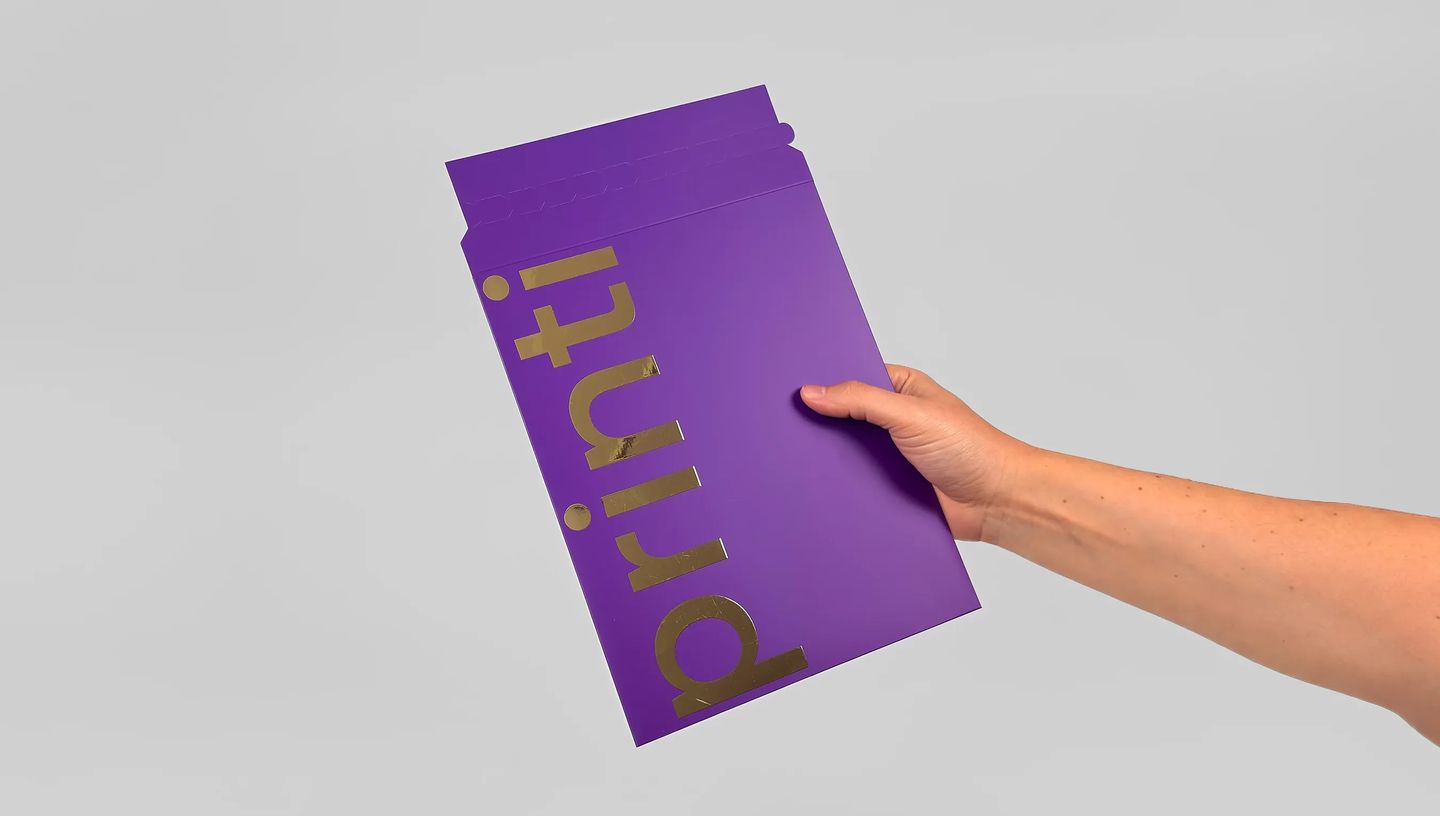 Envelope de segurança personalizado na cor roxa com acabamento em hot stamping dourado exibindo a marca Printi em destaque. O envelope está sendo segurado por uma pessoa e possui aba superior com sistema de fechamento adesivo