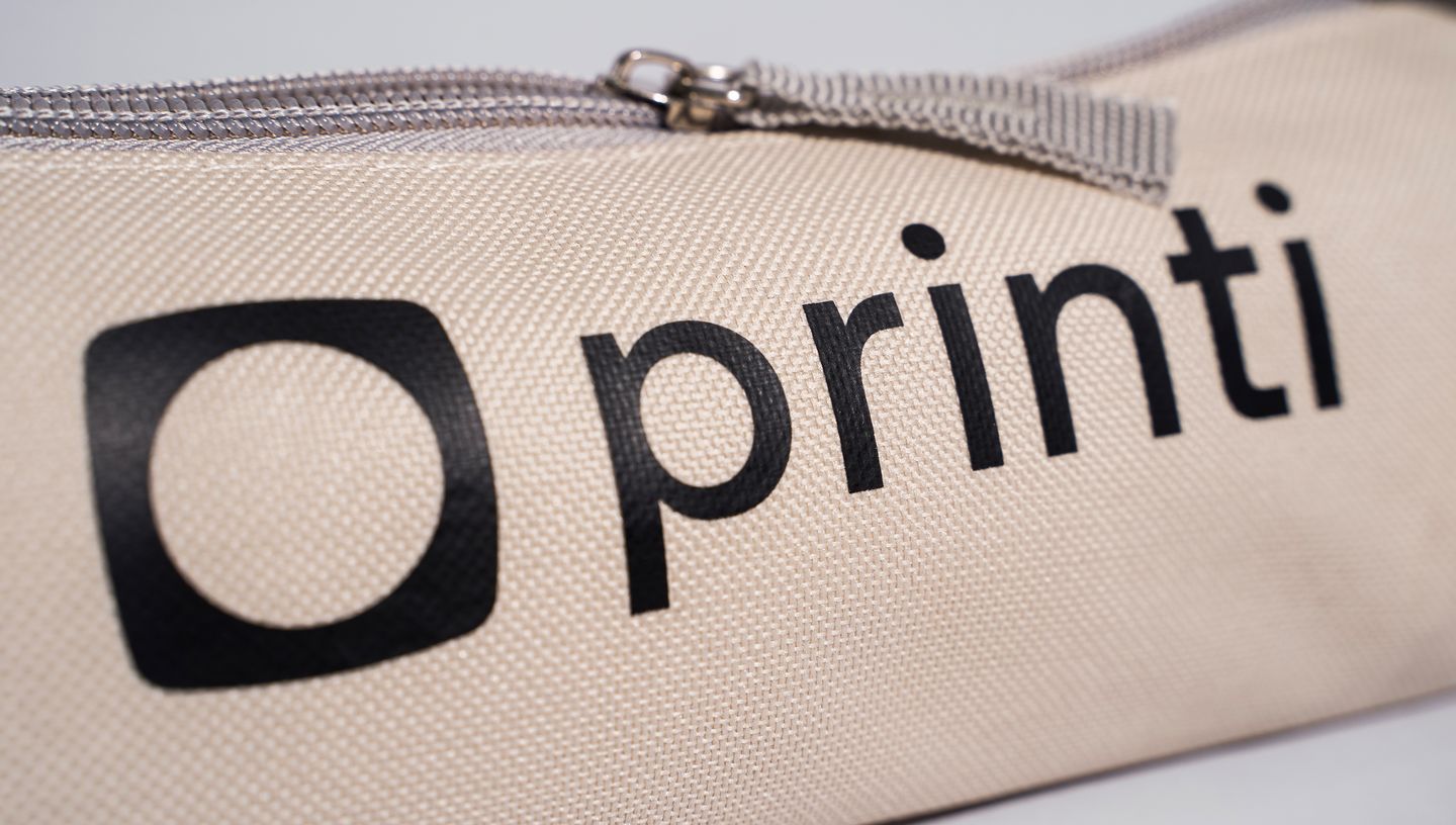 Detalhe do estojo para lápis com tecido resistente em tom bege, zíper metálico e logotipo da Printi estampado em preto