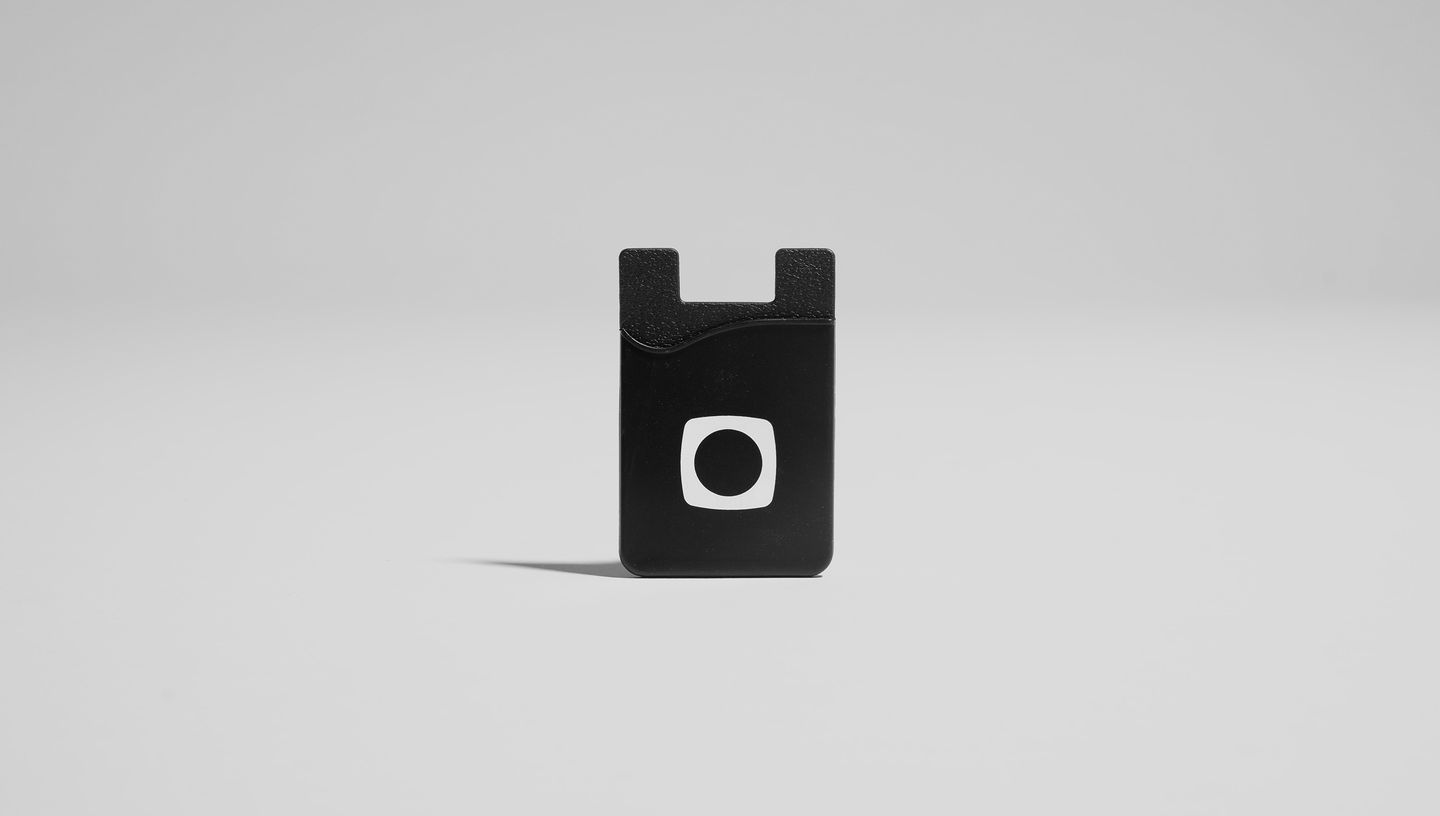 Porta Cartões para Celular em Silicone na cor preta com logotipo branco centralizado, em fundo neutro para destaque do produto