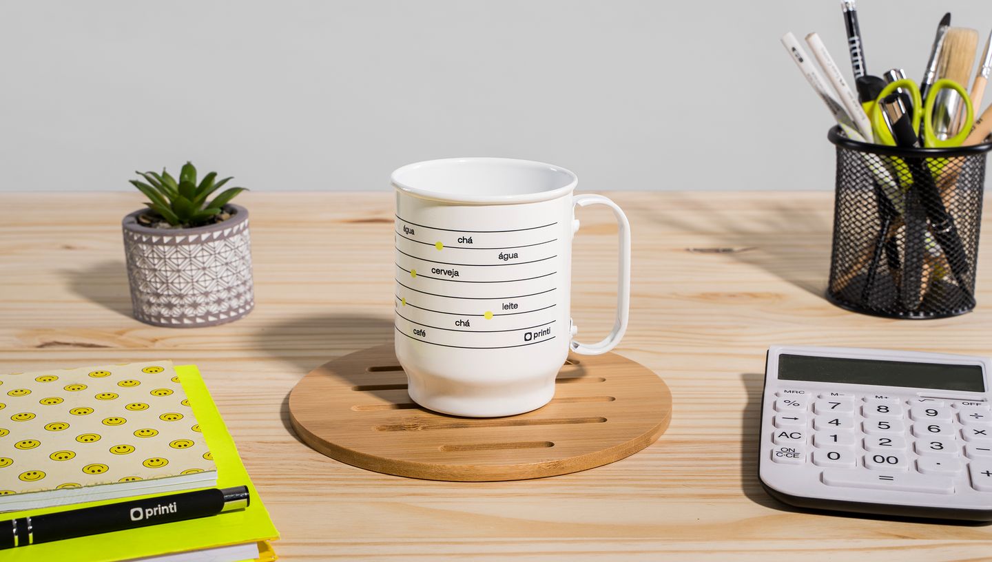 Caneca de alumínio branca apoiada em um suporte de madeira em uma mesa de escritório com itens de decoração ao redor, como calculadora, porta lápis, caderno e vasinho com planta