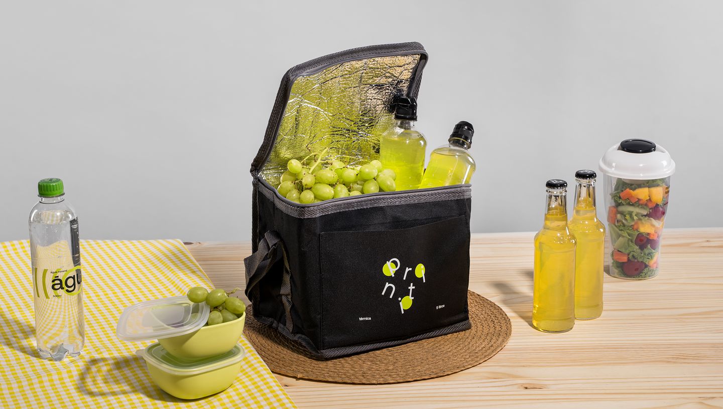 Bolsa térmica preta aberta com bebidas e uvas verdes sobre uma mesa de madeira, acompanhada de garrafas, frutas cortadas e uma garrafa de água.