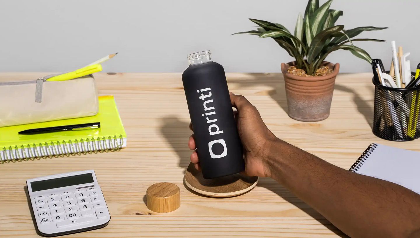 Garrafa squeeze personalizada preta da Printi sendo segurada sobre mesa de trabalho com caderno, calculadora, estojo e planta decorativa ao fundo